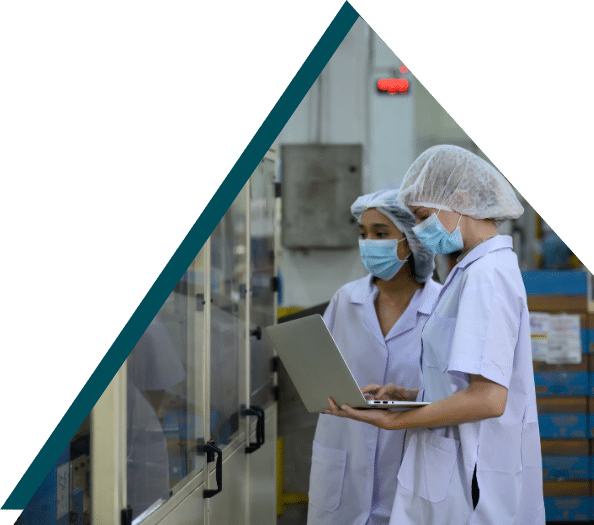 Two healthcare workers in white lab coats and hair nets reviewing data on laptop in sterile food processing facility