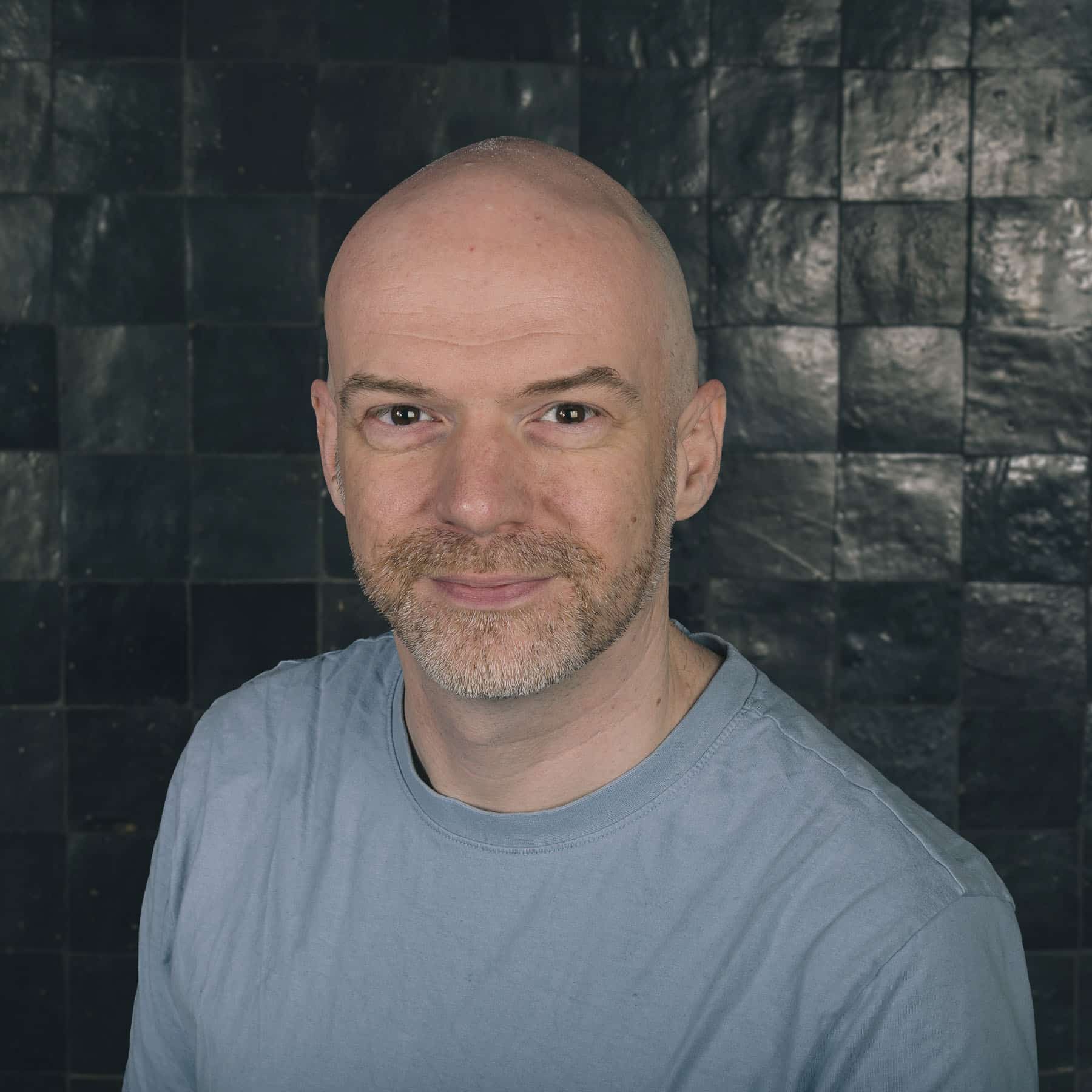 Professional headshot of bald man with beard wearing gray t-shirt against dark tiled background at Sirus tech company
