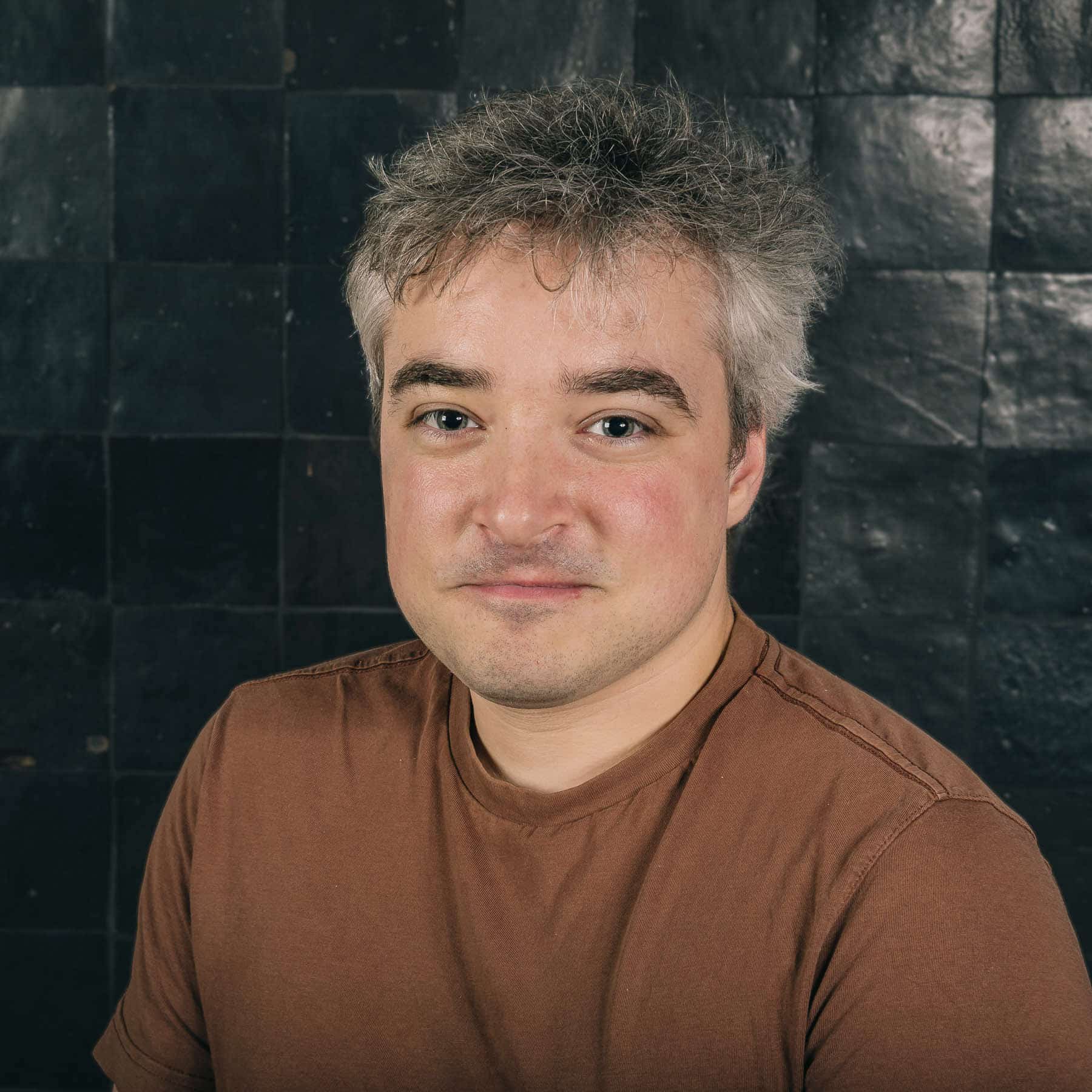 Professional headshot of Stefaan, Sirus team member, smiling in brown shirt against dark brick wall background