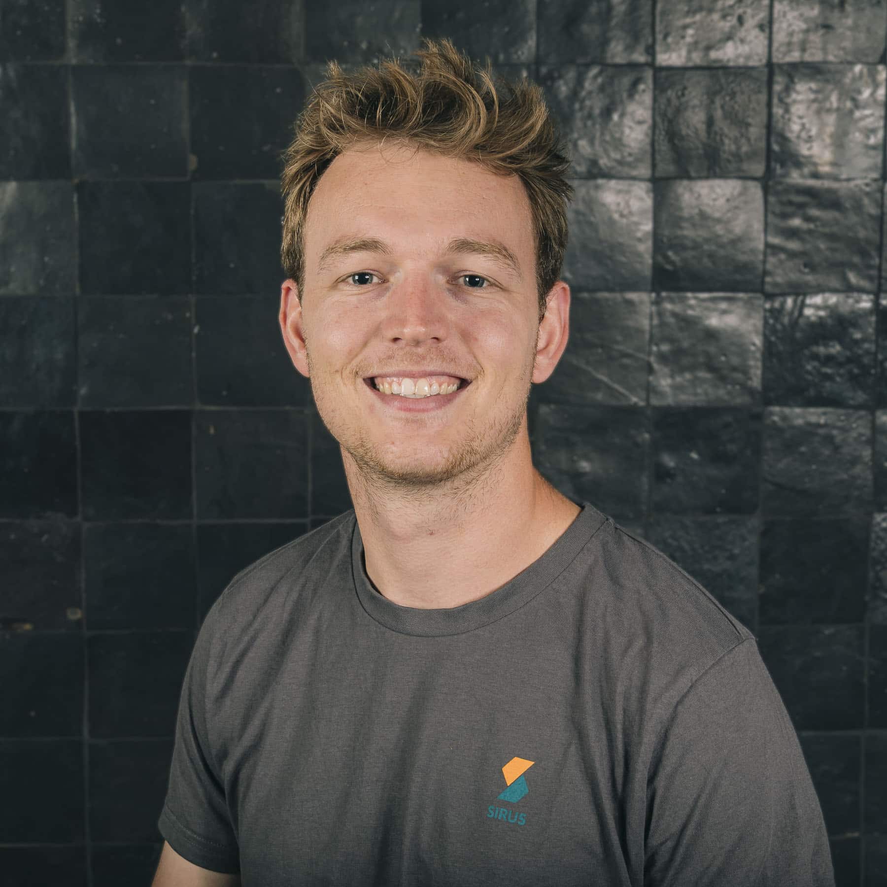 Thomas Devriese smiling in grey Sirus t-shirt against dark tiled wall background