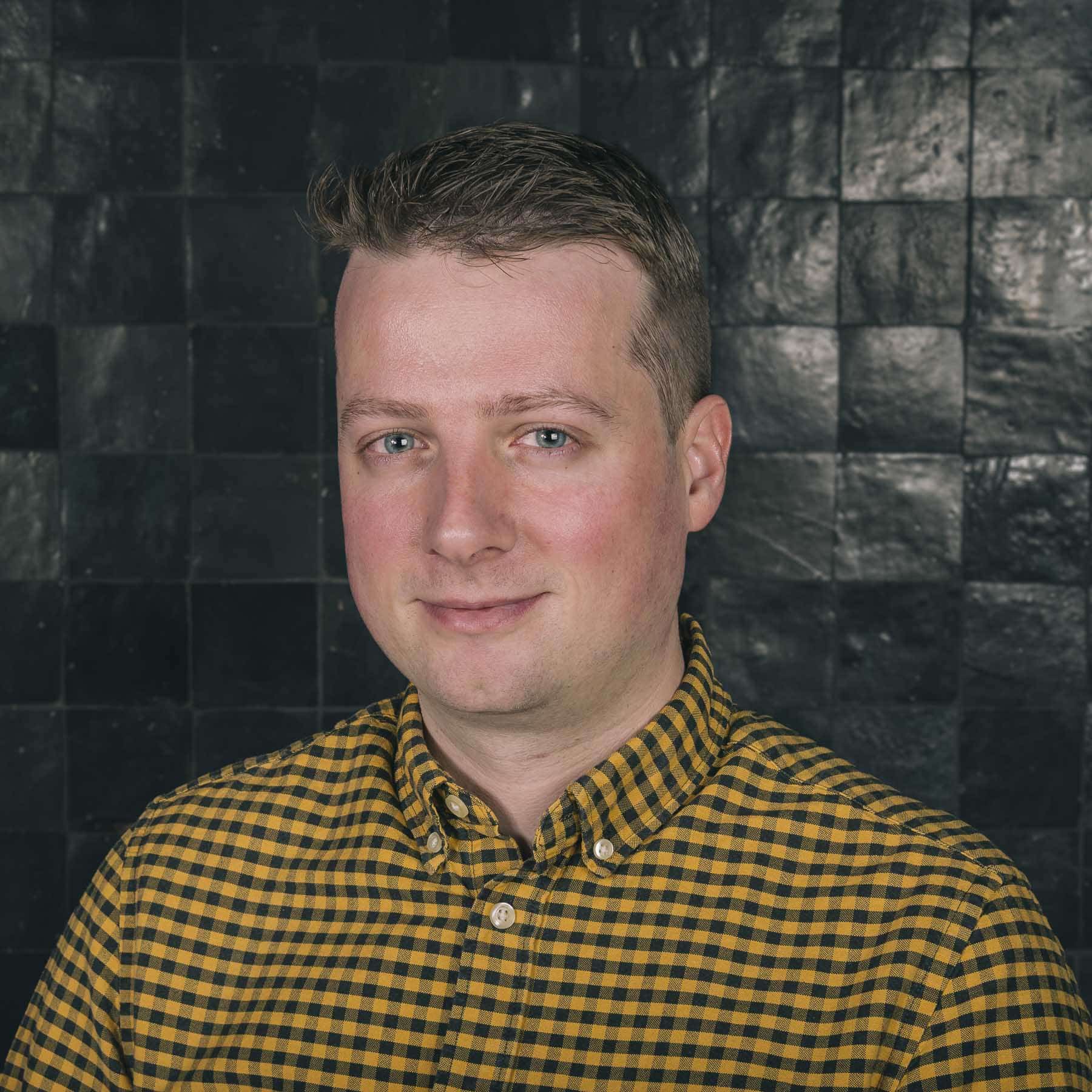 Professional headshot of Timothy, a Sirus team member, wearing a yellow checkered shirt against a dark tiled background