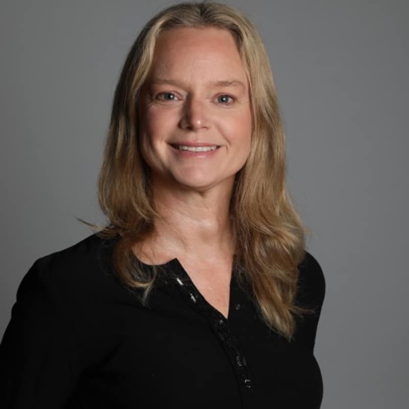 Professional headshot of a smiling blonde woman in a black collared shirt against a gray background