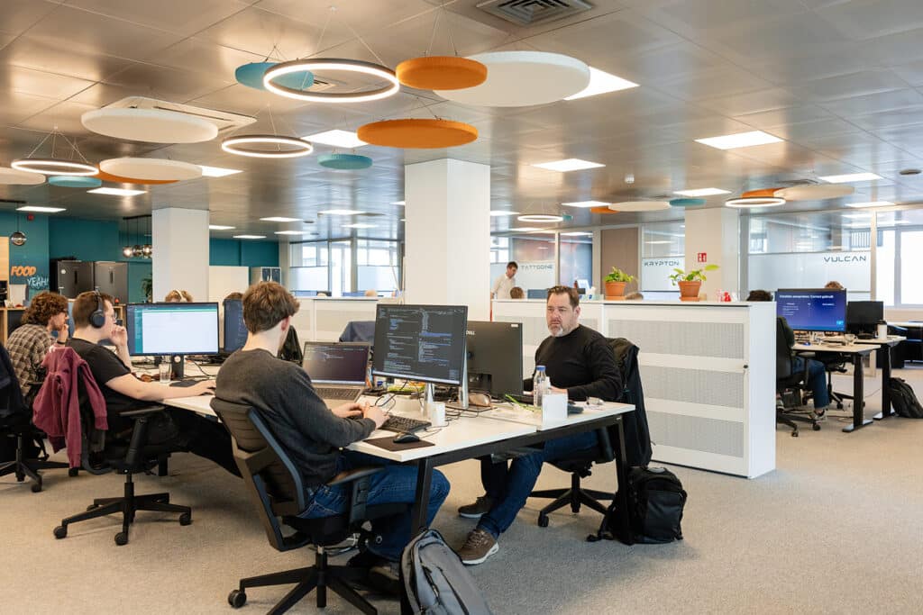 Modern open office with employees working at desks with multiple monitors and colorful suspended ceiling panels