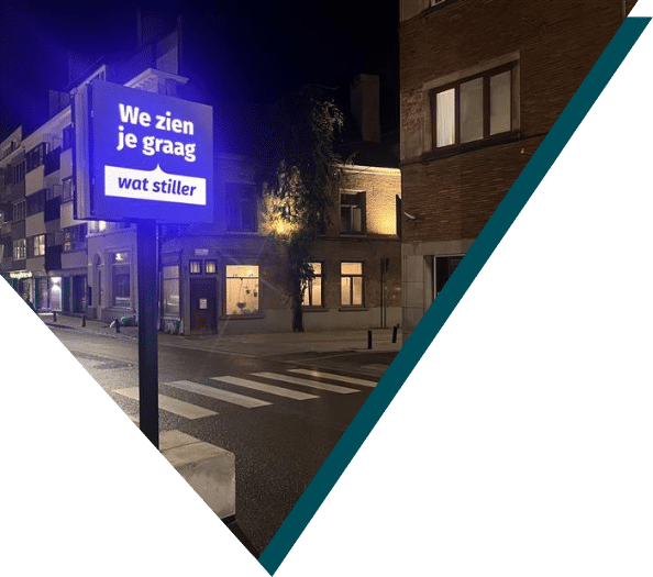 Illuminated blue Smart City sign reading "We zien je graag wat stiller" on a street corner in Ghent, Belgium at night