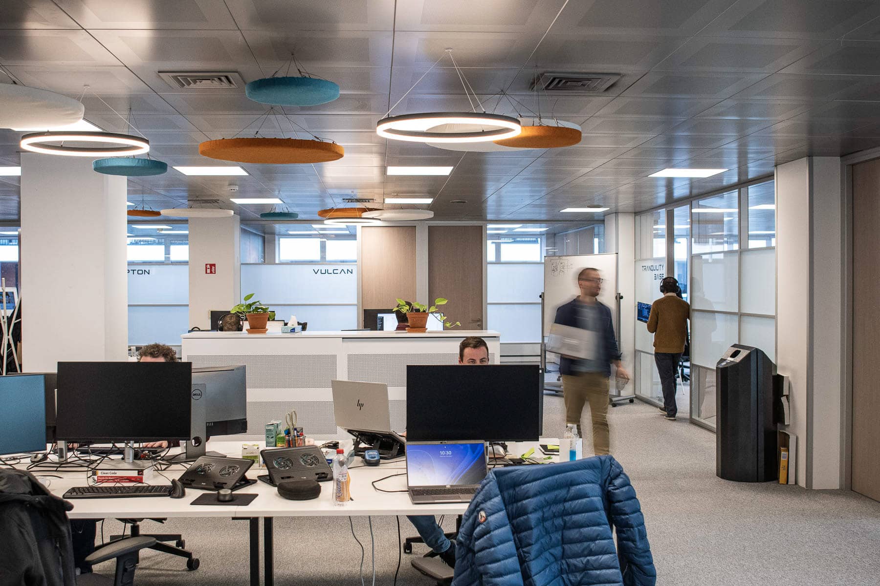 Sirus IT office workspace with developers at desks, monitors, and colorful acoustic ceiling panels in their Merelbeke headqua